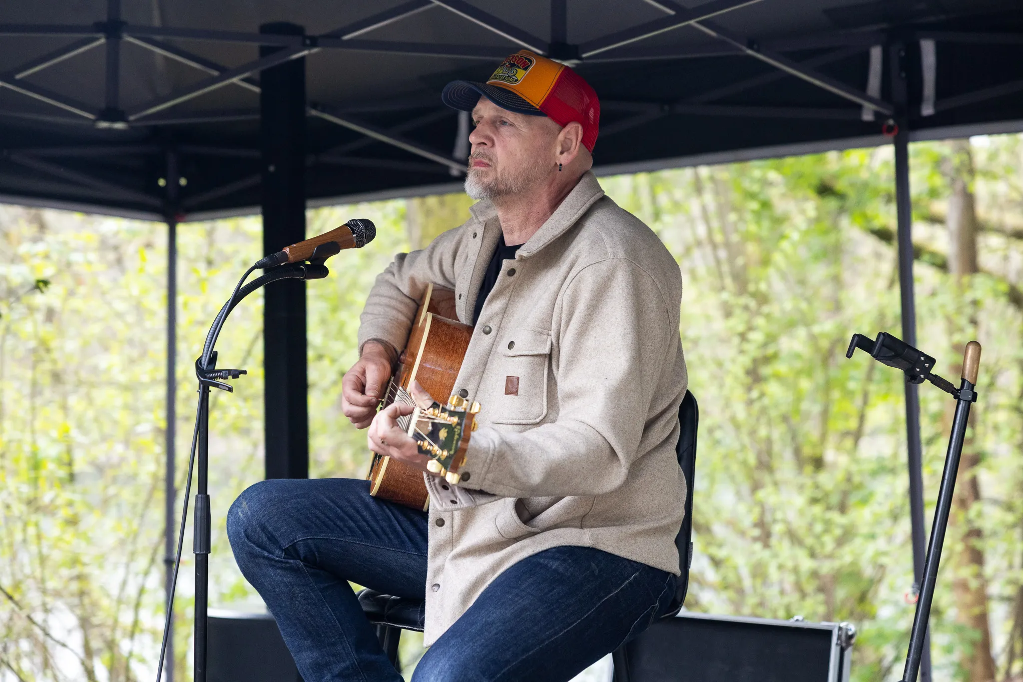 Ein Mann mit grau meliertem Bart spielt auf einer Akustikgitarre, während er auf einem Stuhl sitzt. Er trägt eine Jacke, Jeans und eine Baseballkappe. Im Hintergrund ein Zelt und Bäume.