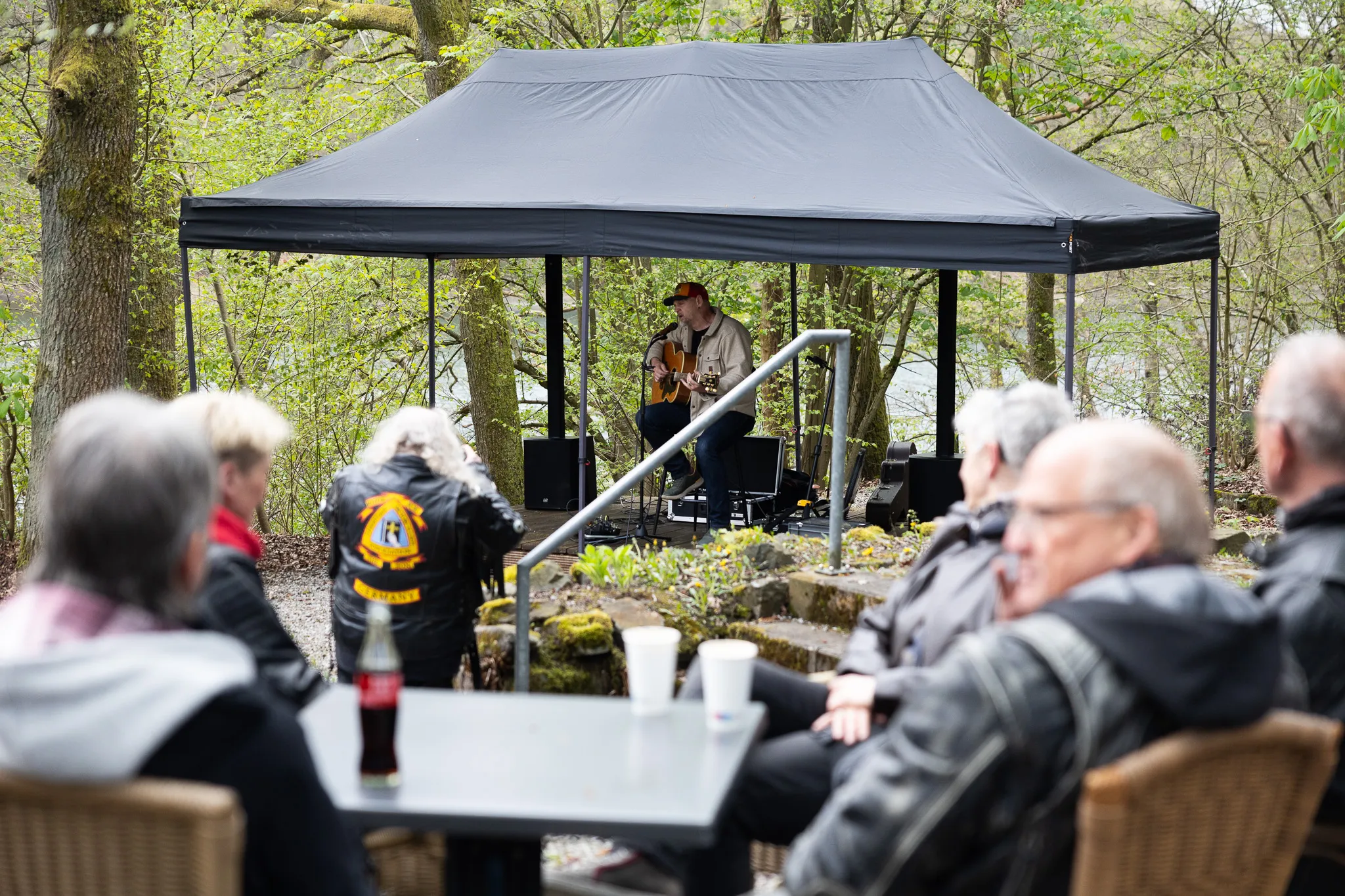 Ein Gitarrist spielt unter einem Pavillon vor einem Publikum aus Motorradfahrern und anderen Zuschauern an einem sonnigen Tag.