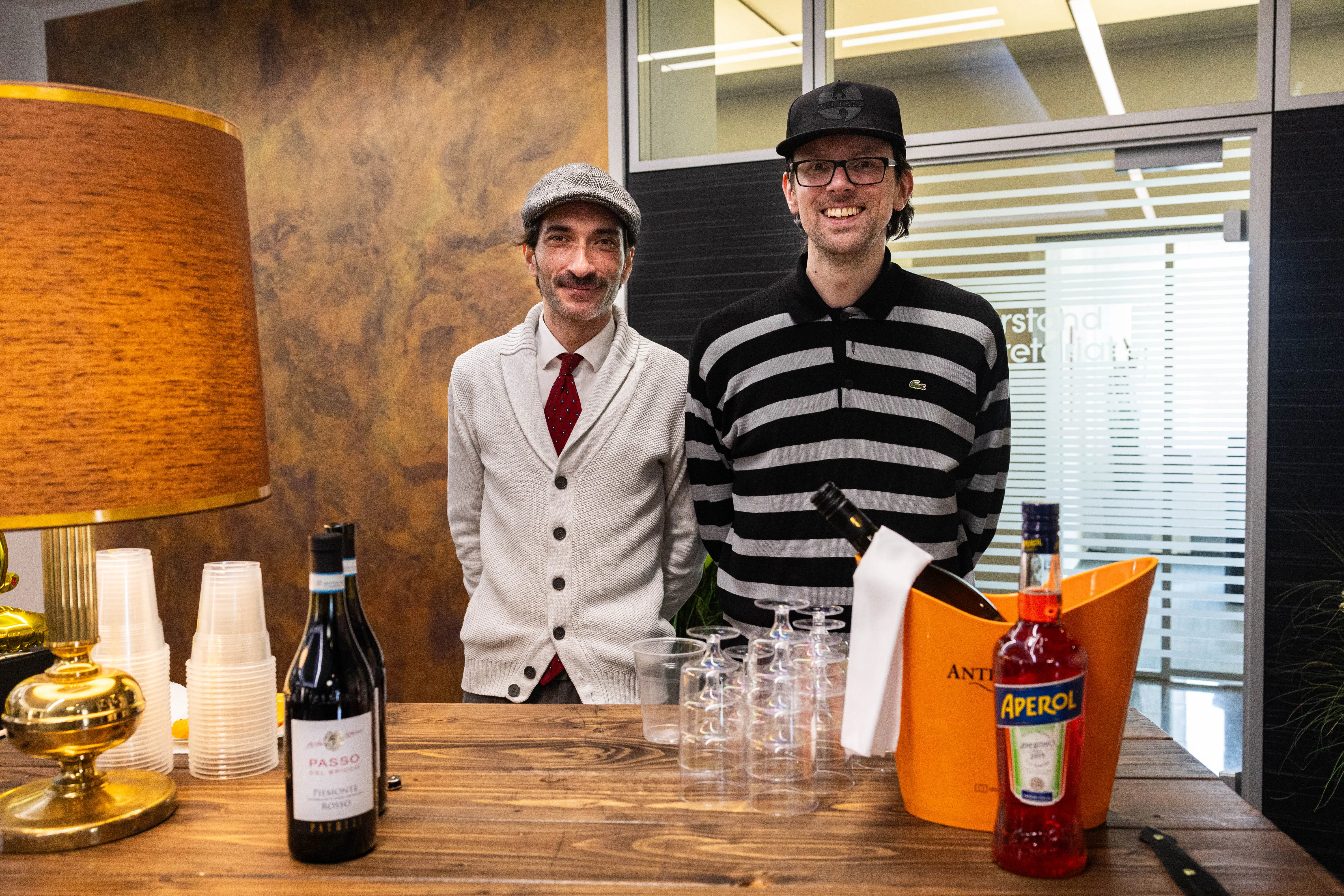 Zwei Männer stehen an einem Tisch mit Getränken und Gläsern, im Hintergrund eine Glastür und ein Büro.