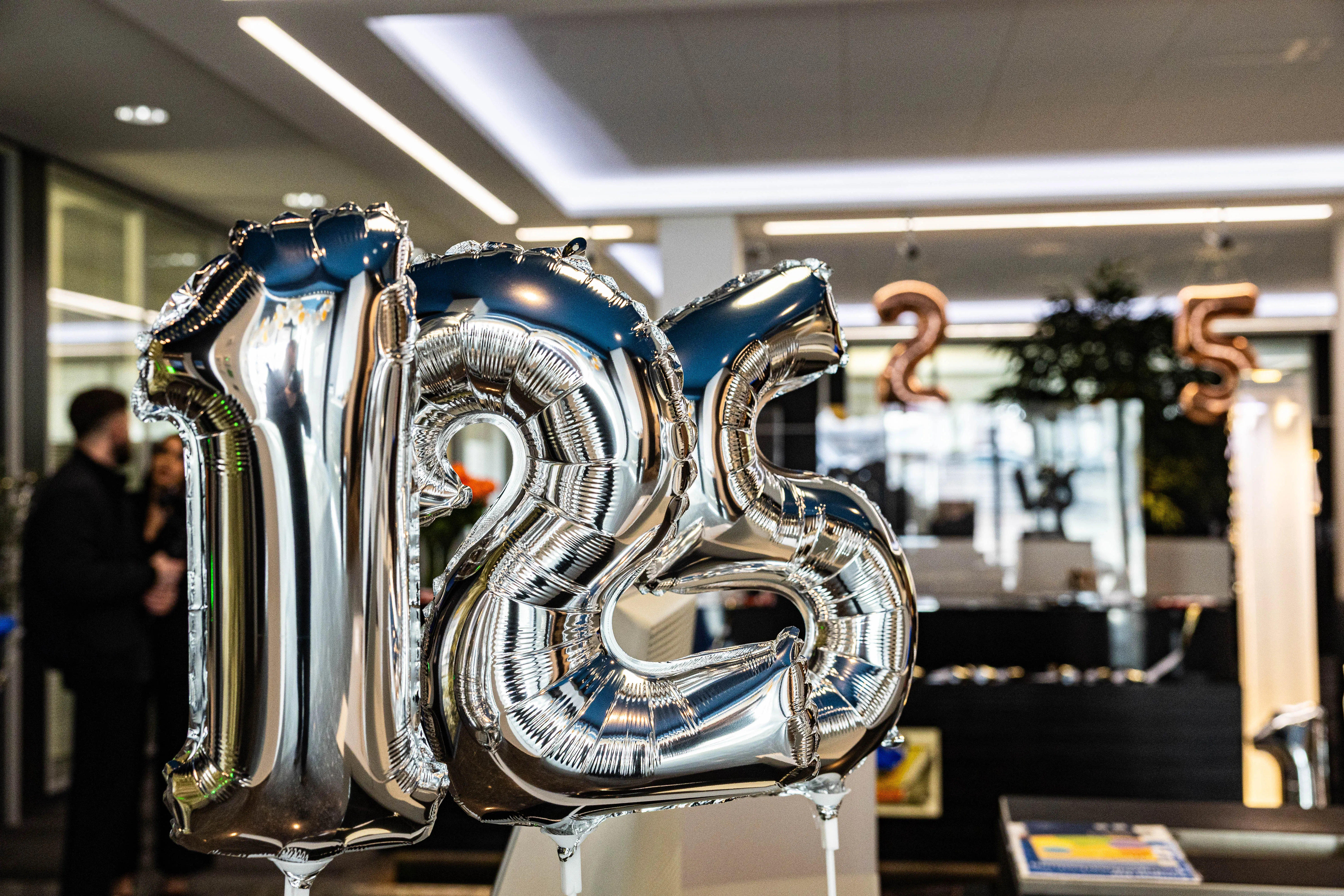 Silberne Folienballons formen die Zahl 125, im Hintergrund verschwommen Menschen bei einer Feier.