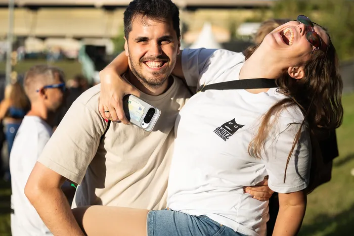 Ein lachender Mann trägt eine lachende Frau im weißen T-Shirt mit Bautz Festival Logo auf einem sonnigen Festivalgelände.