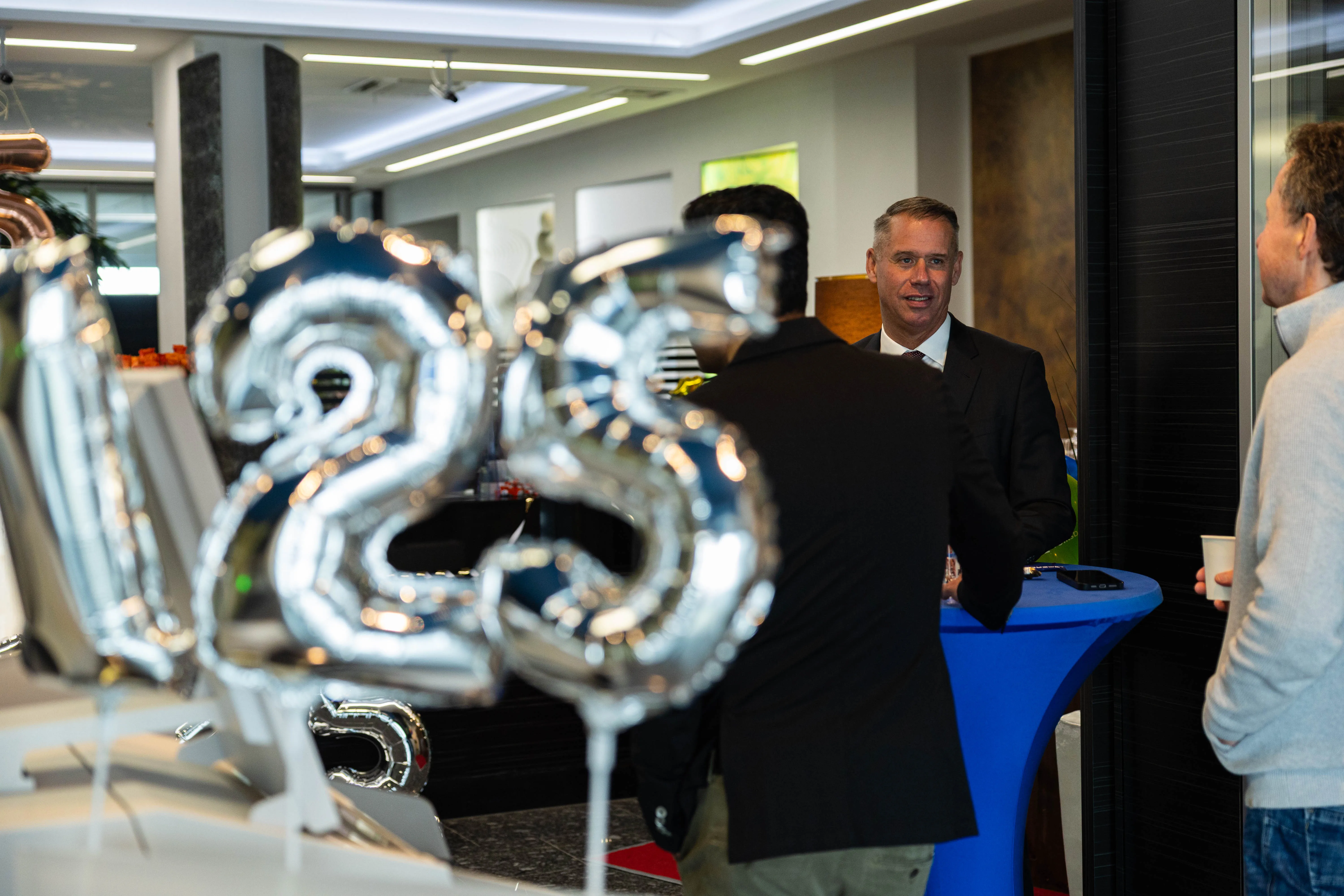 Mann in Anzug lächelt während einer Feier vor silbernen Luftballons mit der Zahl 125. Im Hintergrund ein blauer Empfangstresen und weitere Personen.