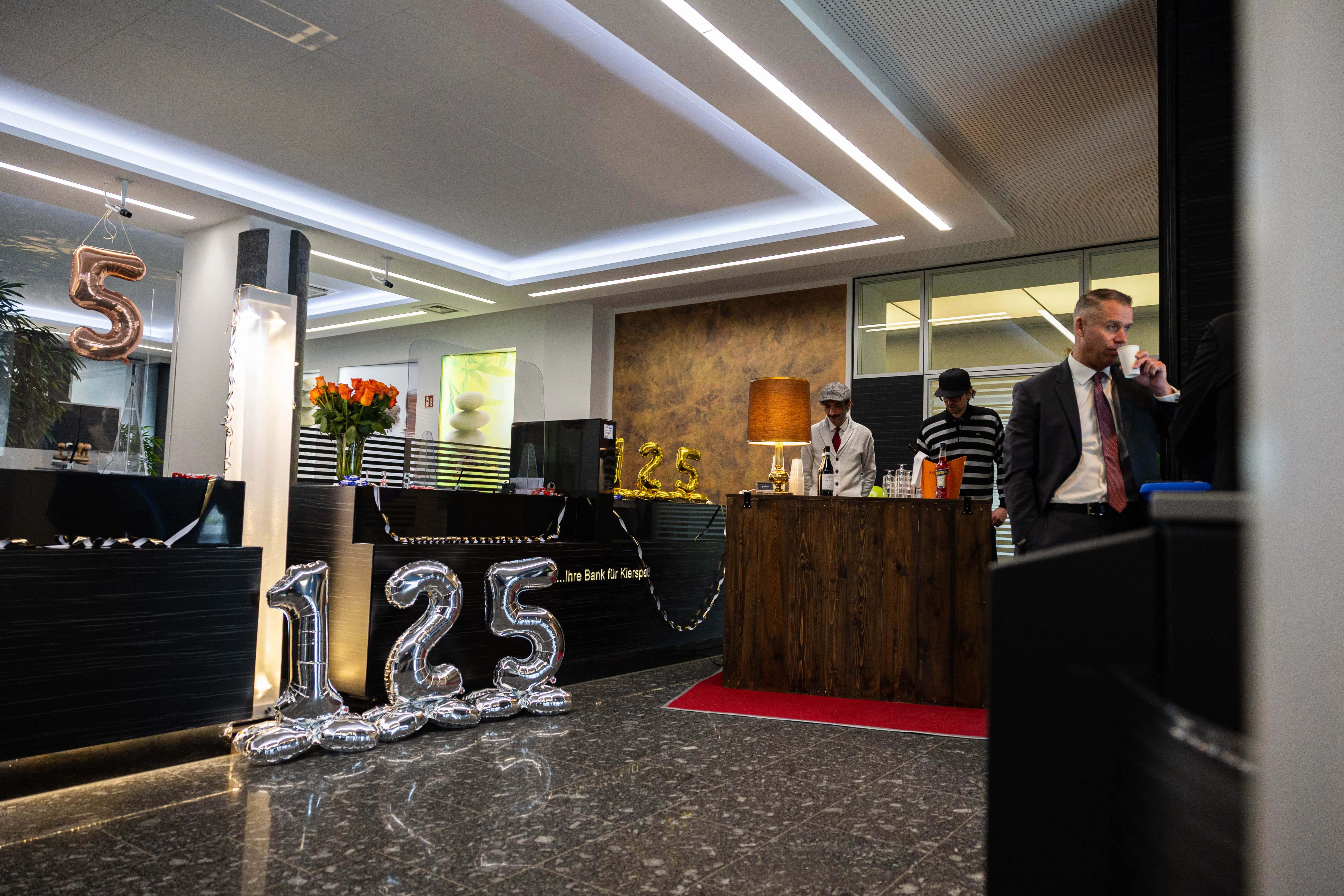 Festlich dekorierter Schalterraum der Volksbank Kierspe mit silbernen Zahlen „125“ und einem Mann mit Anzug, der ein Getränk hält.