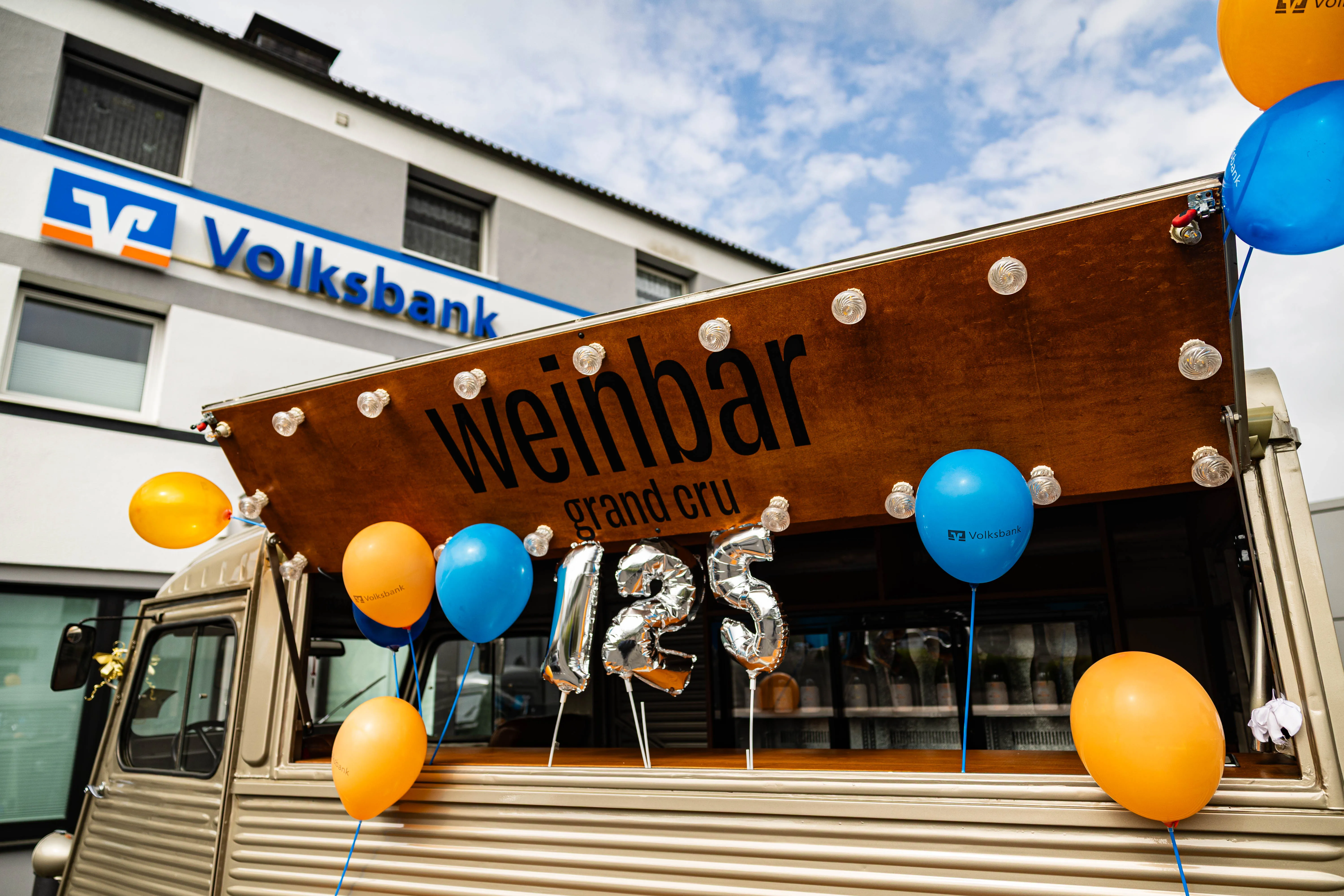 Weißer Foodtruck mit der Aufschrift „Weinbar grand cru“ und silbernen Jubiläumszahlen „125“. Im Hintergrund ein Gebäude der Volksbank.
