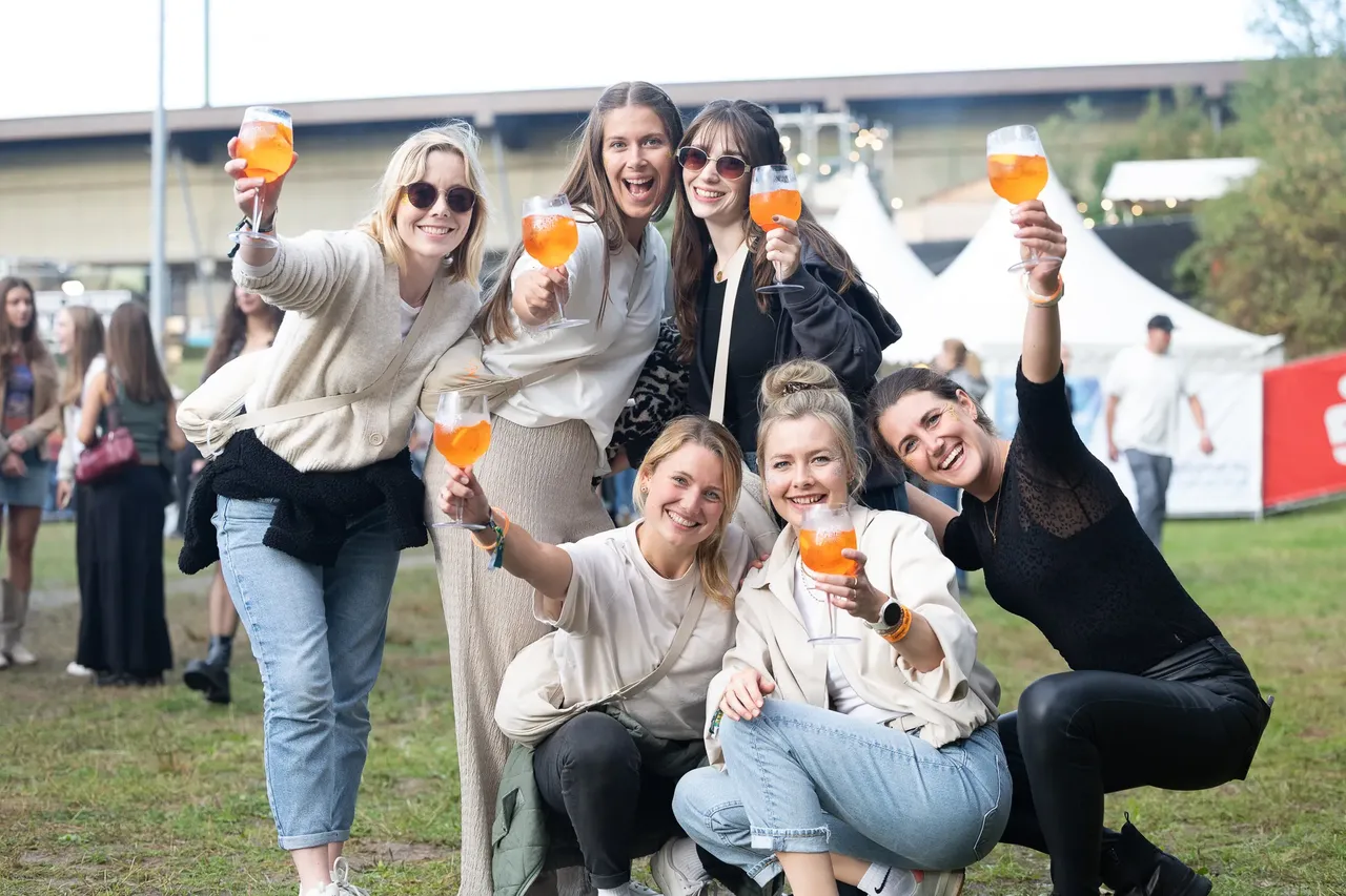 Sechs junge Frauen lächeln und halten Gläser mit orangefarbenen Getränken in die Höhe.