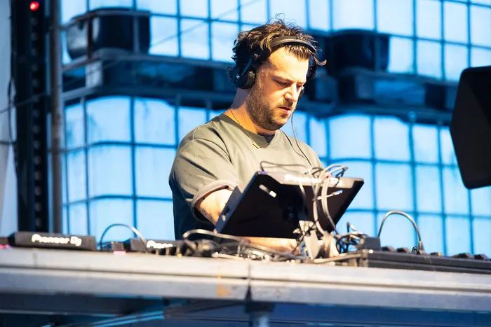 Disco Danny mit Kopfhörern bedient ein DJ-Pult vor einer blau beleuchteten Fensterfront.