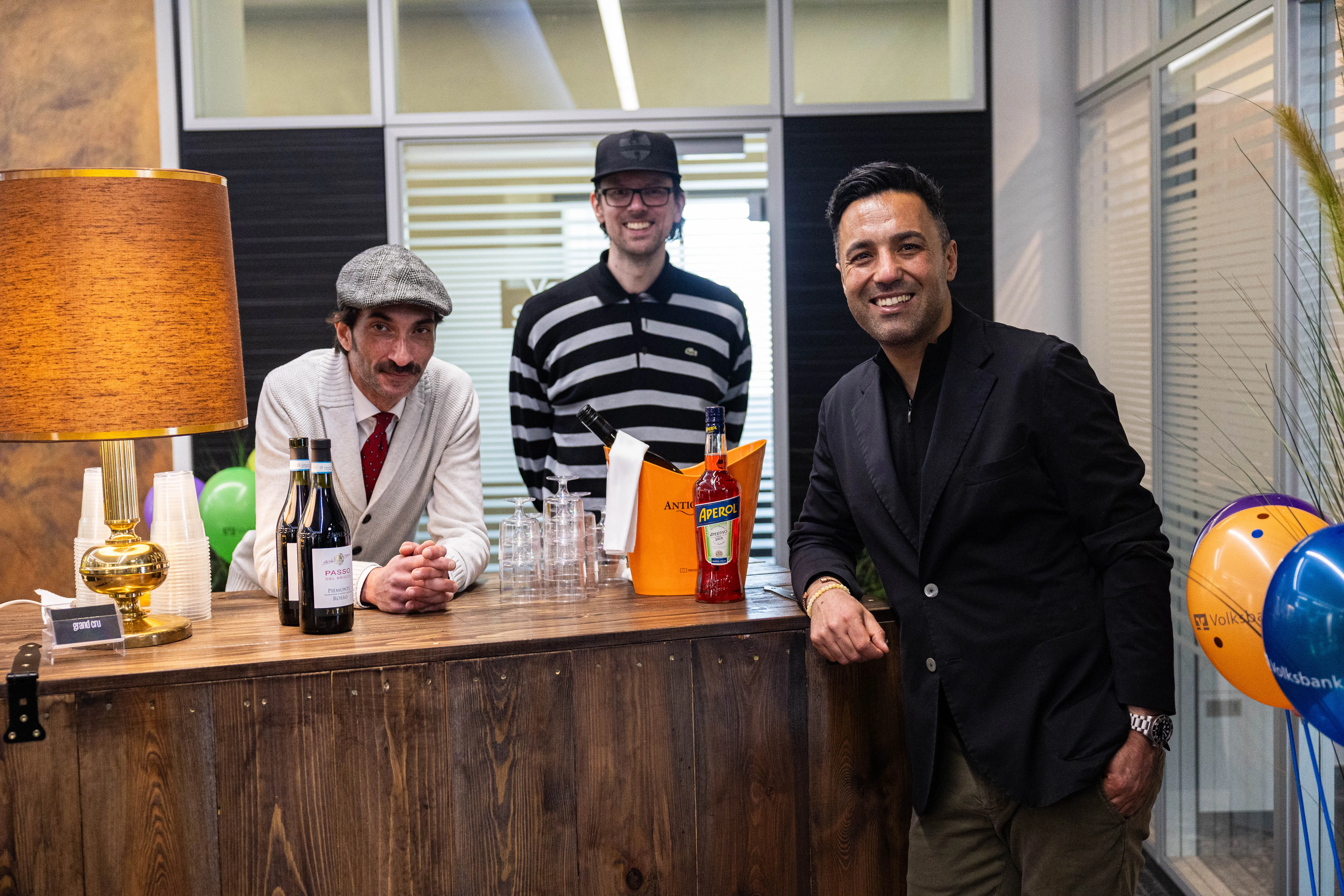 Drei Männer stehen hinter einem Tresen mit Getränken und Weingläsern, lächeln in die Kamera. Im Hintergrund sind Luftballons und ein Volksbank-Logo zu sehen.