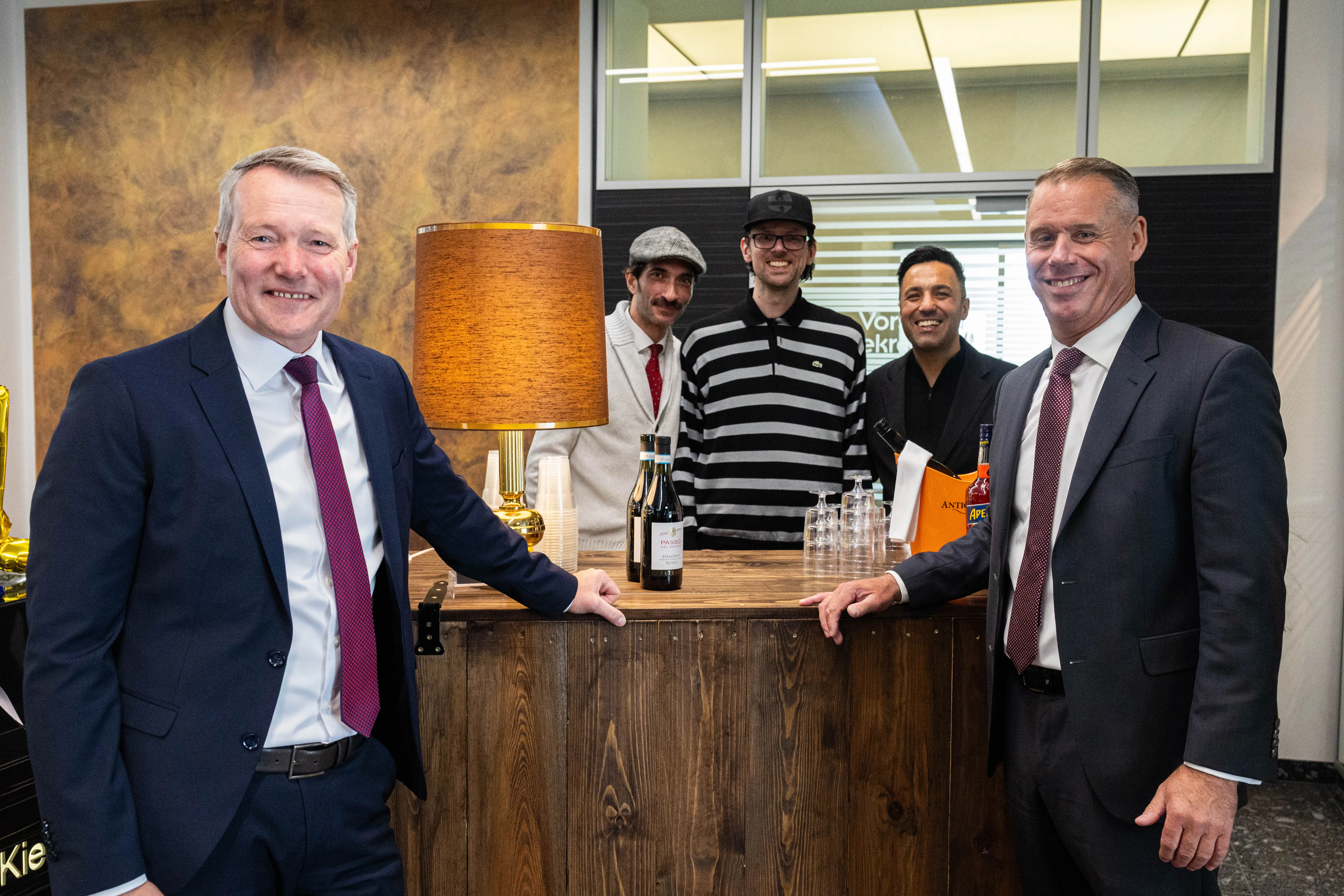 Zwei Männer in Anzügen stehen vor einer Weinbar und reichen sich die Hände. Im Hintergrund stehen drei weitere Personen, darunter Mitarbeiter der Weinbar.