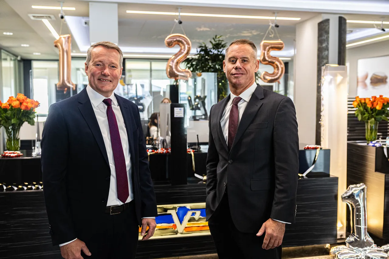 Zwei Männer in Anzug und Krawatte, Stephan Baldschun und Stephan Böhse, stehen vor einer festlich dekorierten Bankfiliale mit silbernen und roségoldenen Jubiläumszahlen.