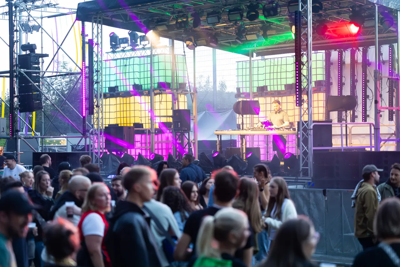 Ein DJ steht an seinem Pult auf einer bunt beleuchteten Bühne mit leuchtenden Paneelen vor einer Menschenmenge.