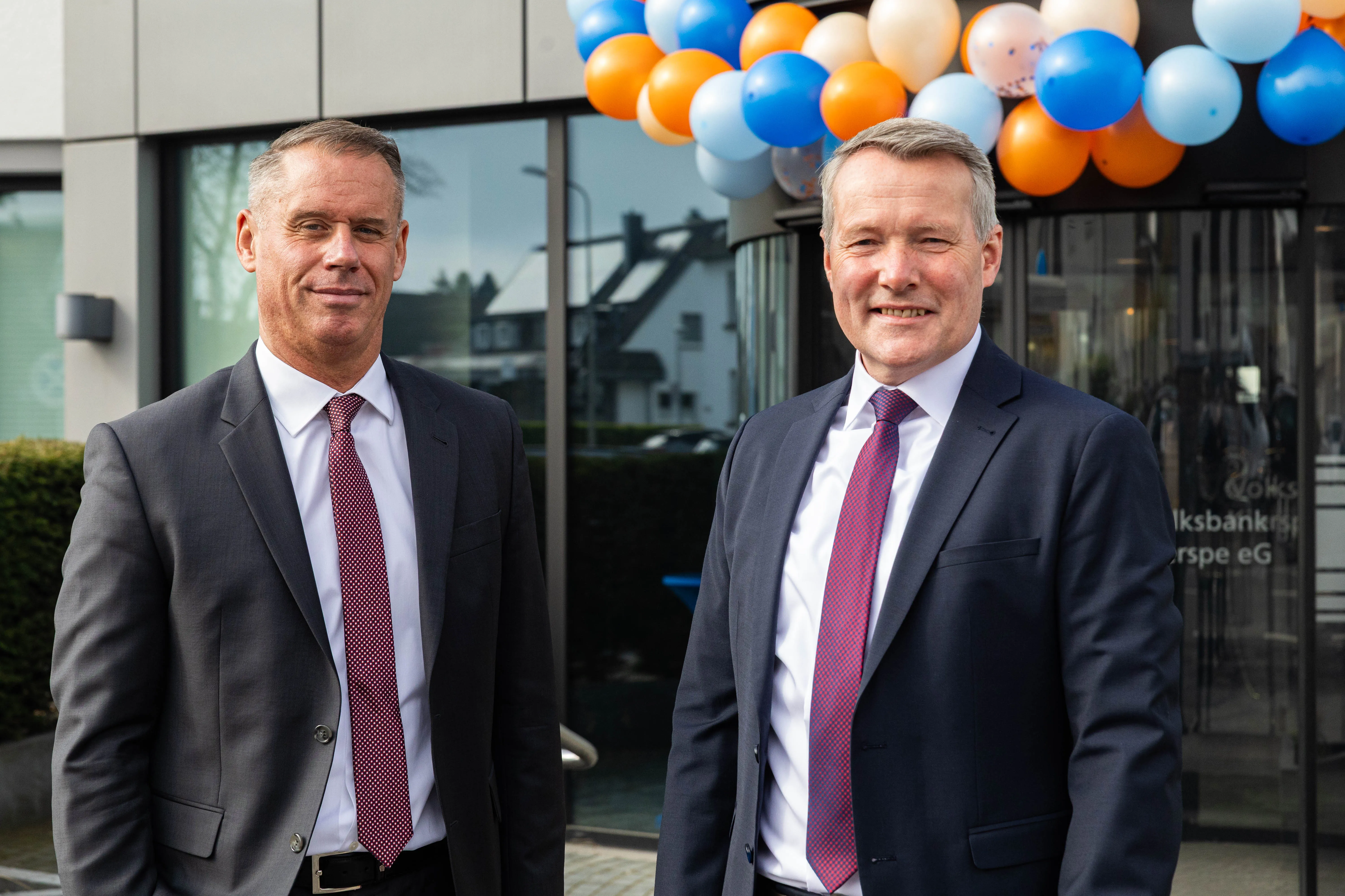 Zwei Männer in Anzug und Krawatte stehen vor dem Eingang einer Bank, im Hintergrund Luftballons in Orange und Blau. Es sind die beiden Vorstände der Volksbank Kierspe, Stephan Baldschun und Stephan Böhse.