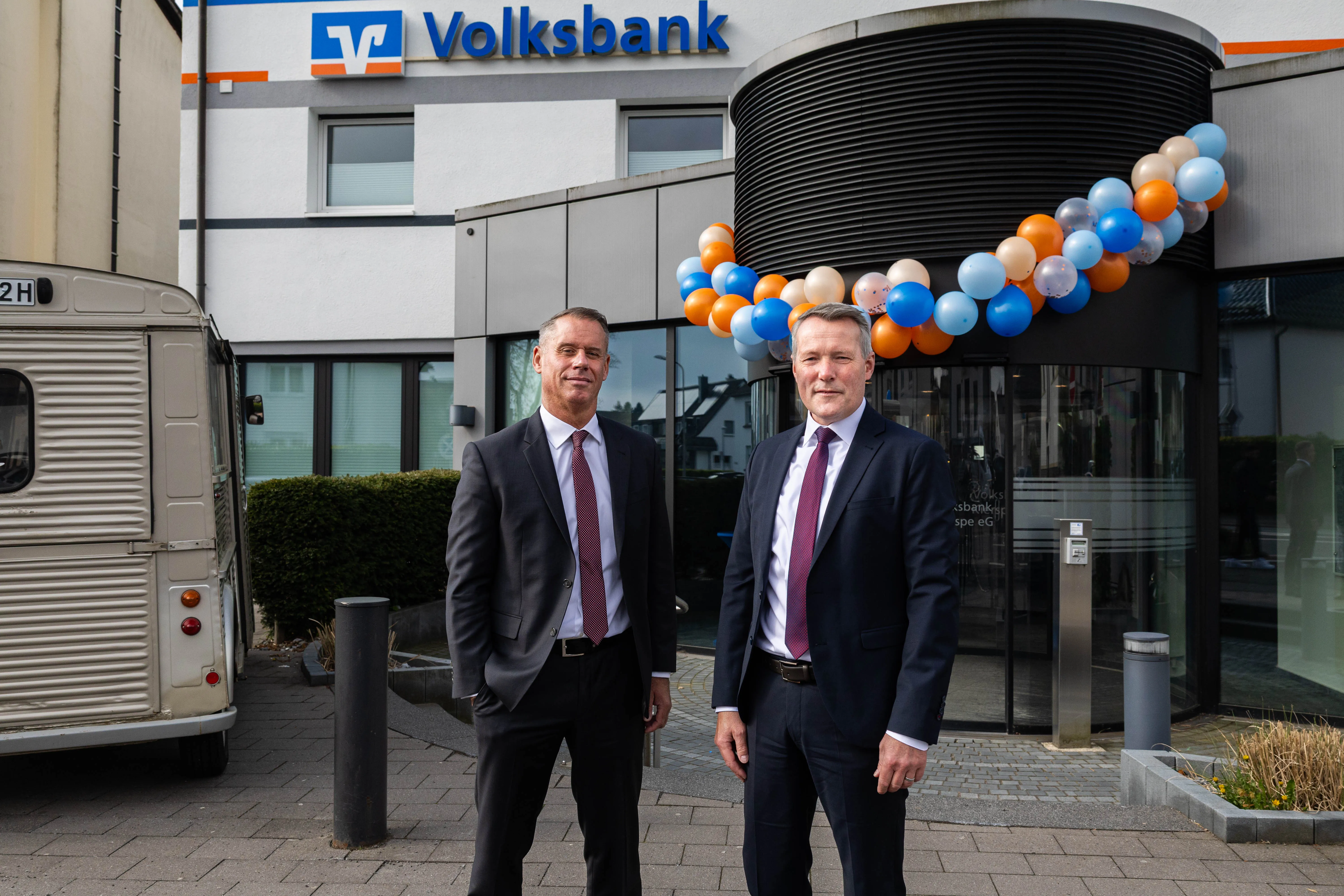 Zwei Männer in Anzug und Krawatte stehen vor der Volksbank Kierspe, die mit bunten Luftballons geschmückt ist. Im Hintergrund ein Foodtruck.