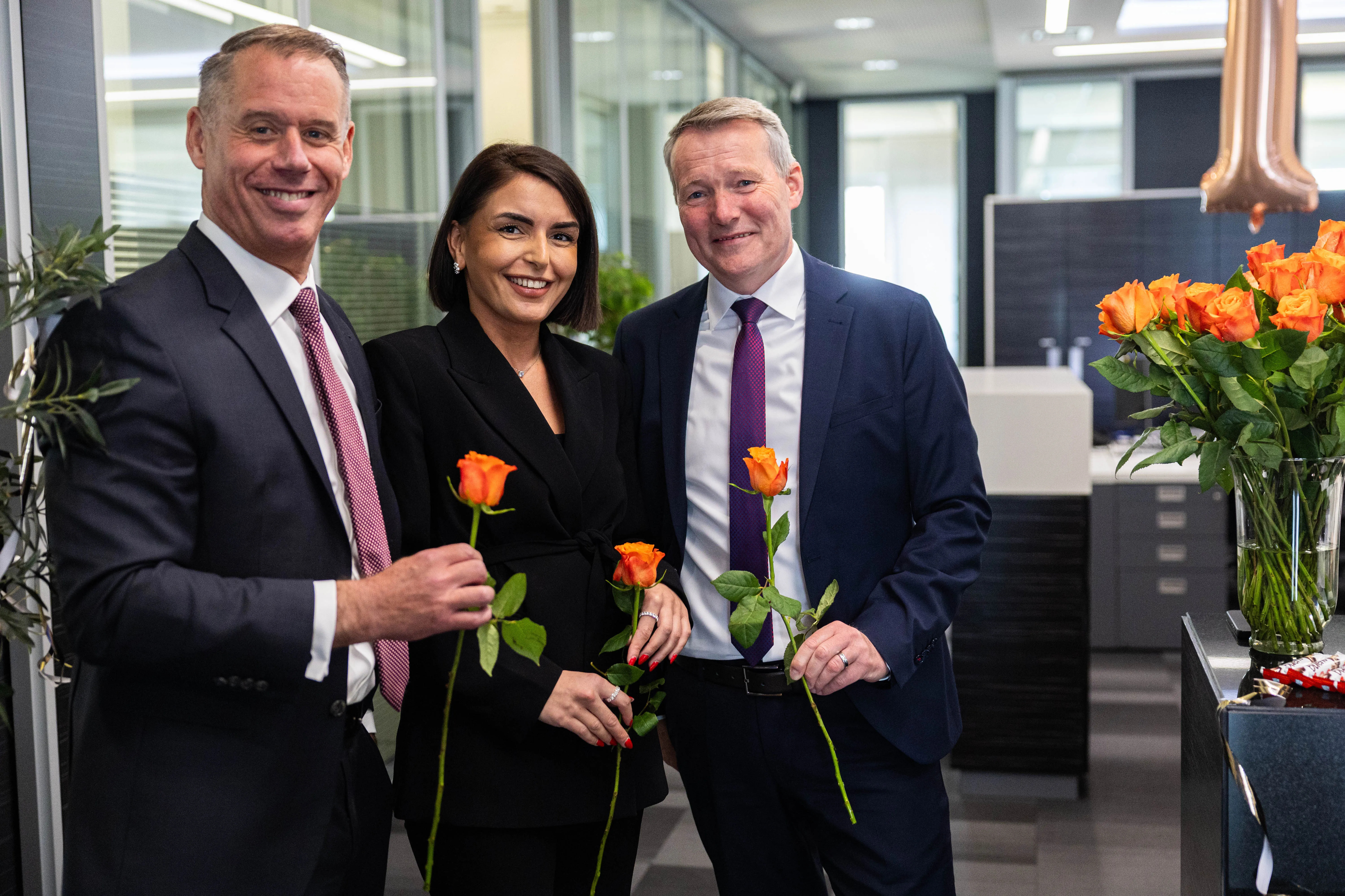 Zwei Männer und eine Frau in formeller Kleidung halten jeweils eine orangefarbene Rose. Sie stehen vor einer modernen Büroumgebung und feiern das 125-jährige Jubiläum der Volksbank Kierspe.