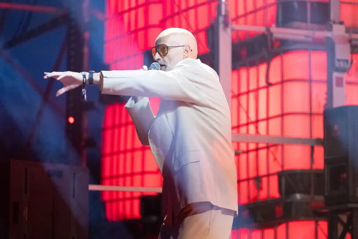 Mambo Kurt im hellen Anzug singt mit Sonnenbrille in ein Mikrofon vor einer rot beleuchteten Bühnenkonstruktion.