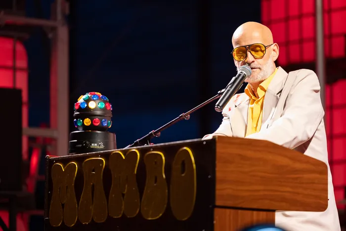 Mambo Kurt mit gelber Sonnenbrille singt in ein Mikrofon hinter einem Pult mit der Aufschrift „MAMBO“, auf dem eine bunte Discokugel-Leuchte steht.