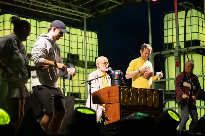 Mambo Kurt steht hinter einem DJ-Pult mit der Aufschrift „MAMBO“ vor einer Kulisse aus grün leuchtenden Paneelen.