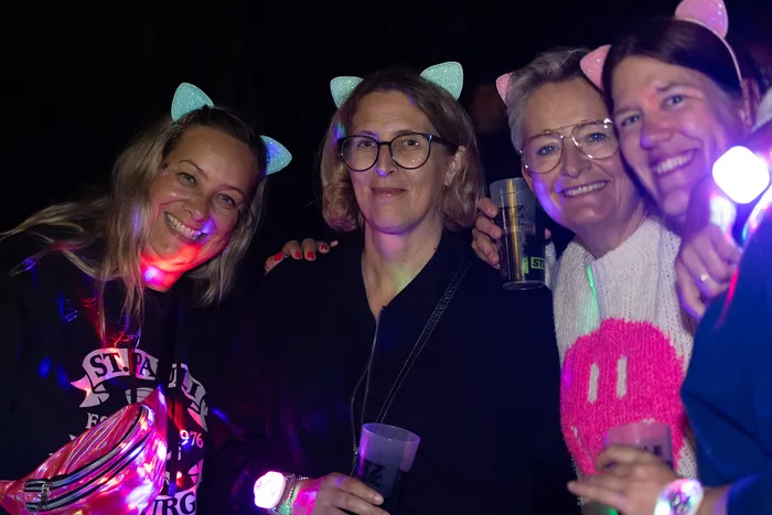 Vier lächelnde Frauen mit Katzenohren-Haarreifen halten Getränke bei einem nächtlichen Event mit bunten Lichteffekten.