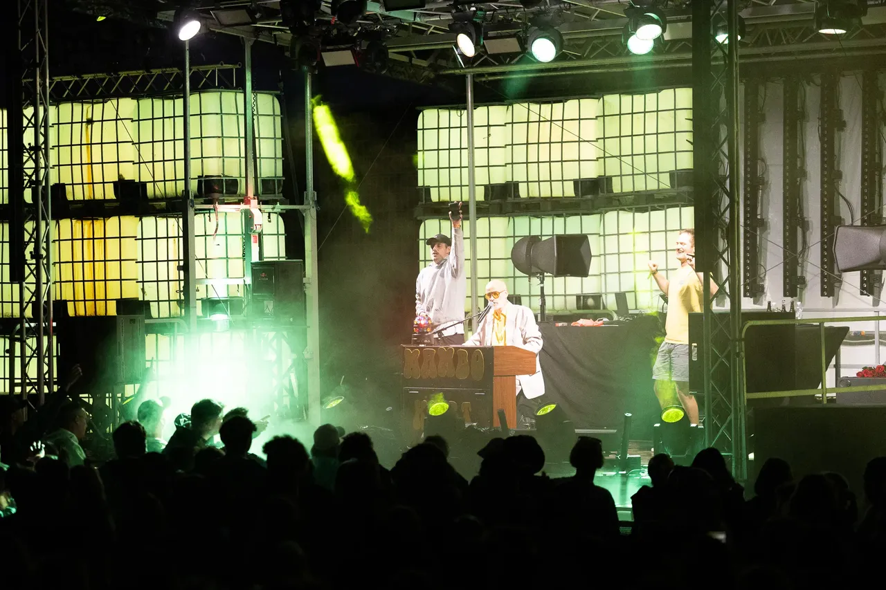 Mambo Kurt und Der Wolf performen an einem DJ-Pult mit der Aufschrift „MAMBO“ unter grünem Bühnenlicht vor einem Publikum.