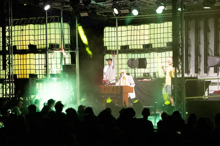 Mambo Kurt und Der Wolf performen an einem DJ-Pult mit der Aufschrift „MAMBO“ unter grünem Bühnenlicht vor einem Publikum.