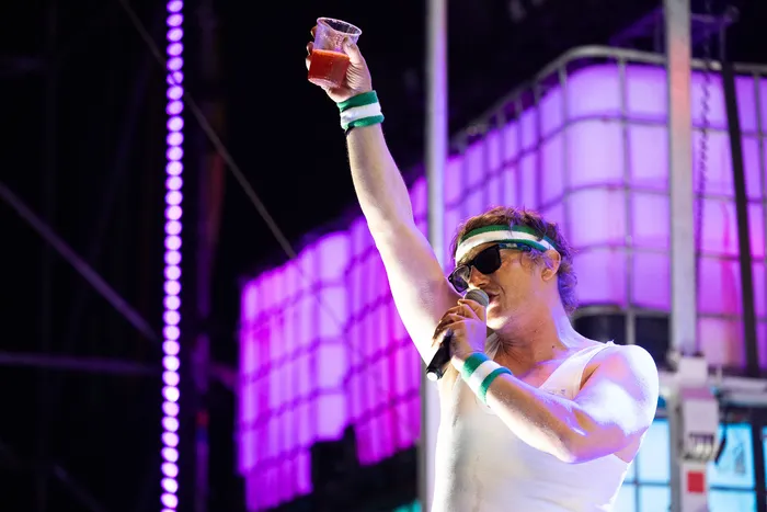 Ein Mann mit Sonnenbrille und weißem Tanktop singt in ein Mikrofon und hält ein Glas mit roter Flüssigkeit hoch. Er trägt grün-weiße Armbänder vor einer lila Bühne.