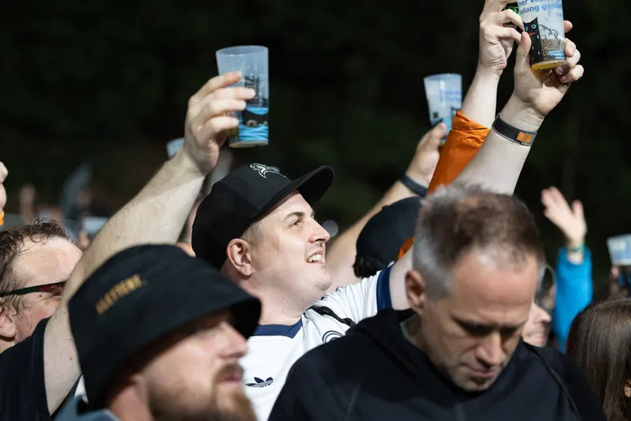 Ein lächelnder Mann mit schwarzer Kappe hebt einen Plastikbecher in die Höhe, während Menschen in der Menge ebenfalls Becher hochhalten.