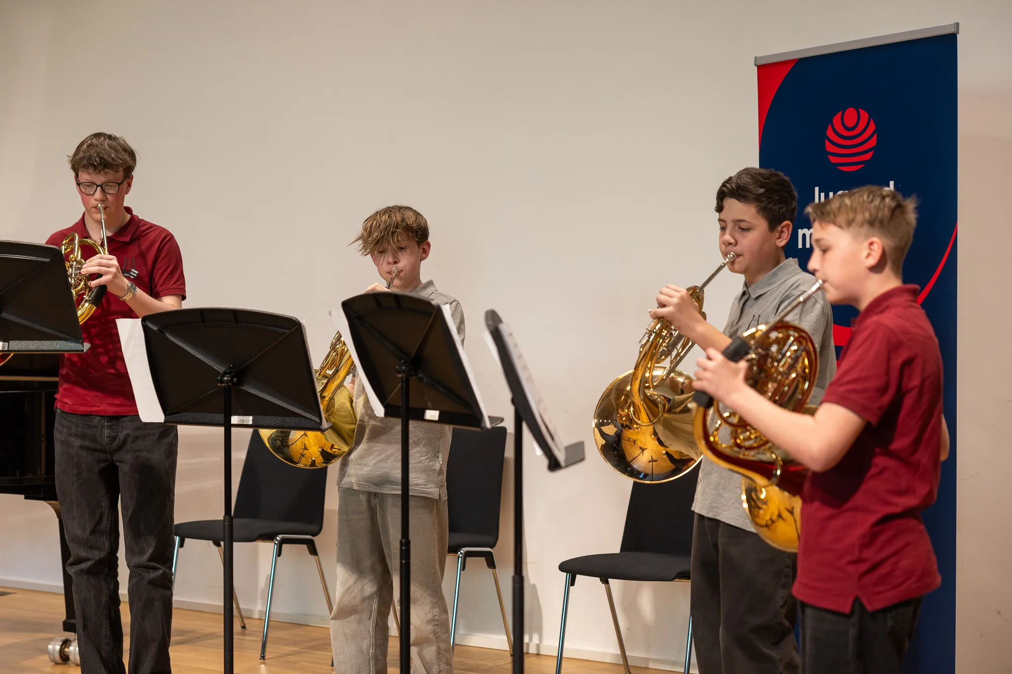 Vier Jungen spielen auf der Bühne Blasinstrumente: Saxophon, Posaune, Euphonium und Tuba. Im Hintergrund steht ein Banner und Notenständer.