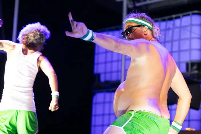 Zwei Performer in grüner Sportkleidung führen eine Tanzperformance auf einer blau beleuchteten Bühne auf.