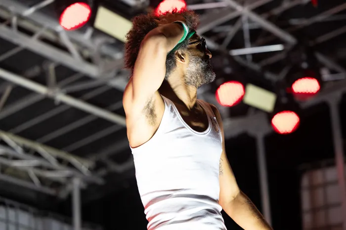 Ein bärtiger Mann mit Afro und weißem Tanktop hebt den Arm an den Kopf vor roten Bühnenlichtern.