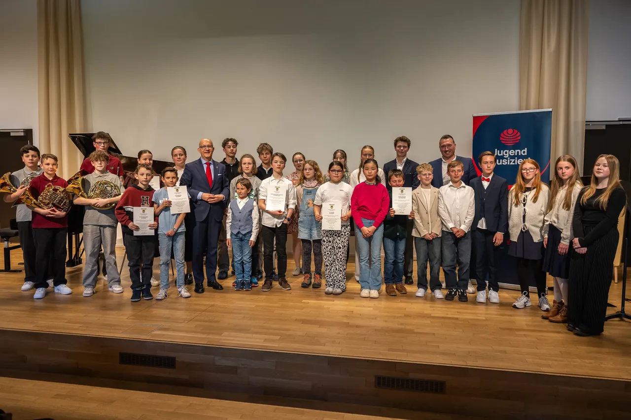 Gruppe von Kindern und Jugendlichen mit Urkunden auf einer Bühne, ein Mann in Anzug steht zwischen ihnen. Im Hintergrund ein Banner mit der Aufschrift „Jugend musiziert“.