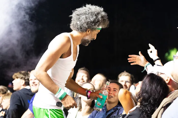 Ein Mann mit Afro-Frisur und weißem Tanktop interagiert bei Nacht mit einer Menschenmenge und hält eine Flasche.