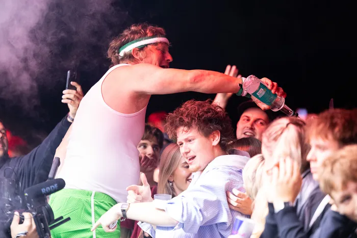 Ein Mann in weißem Tanktop und grünem Stirnband hält eine Flasche über eine Menschenmenge auf der Turntable Stage.