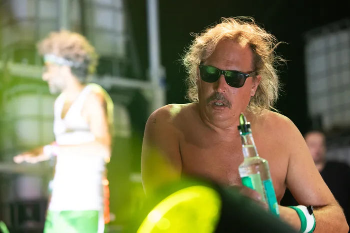 Ein oberkörperfreier Mann mit langen blonden Haaren und Sonnenbrille hält eine Flasche mit blauer Flüssigkeit auf der Turntable Stage.