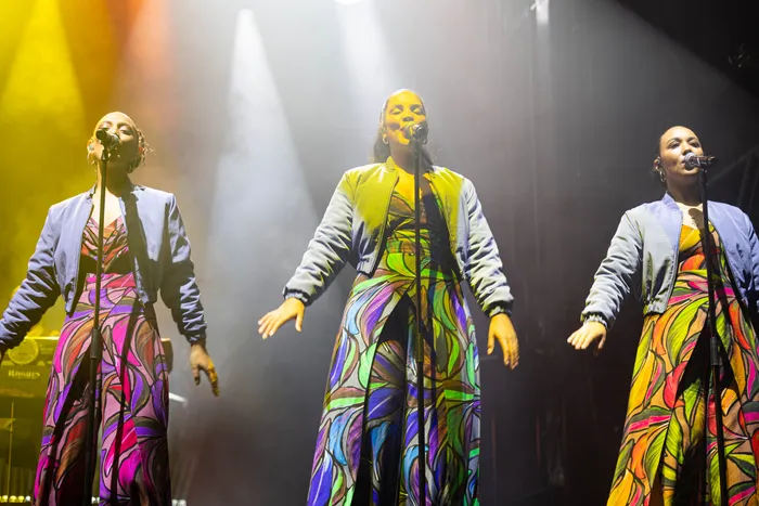 Drei Sängerinnen in farbenfrohen, gemusterten Outfits und hellblauen Jacken singen auf einer Bühne unter Scheinwerferlicht.