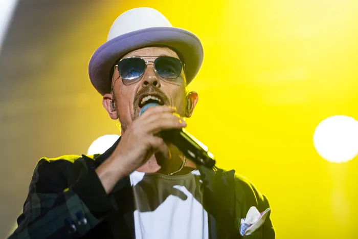 Jan Delay mit hellblauem Hut und Sonnenbrille singt in ein Mikrofon vor gelbem Bühnenlicht.