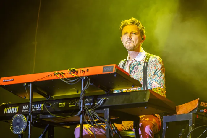 Ein Musiker mit bunt gemustertem Hemd und Hosenträgern spielt an Keyboards auf einer Bühne unter gelbem Licht.