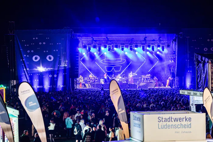 Musiker auf einer blau beleuchteten Bühne vor einer großen Menschenmenge bei einem nächtlichen Open-Air-Konzert.