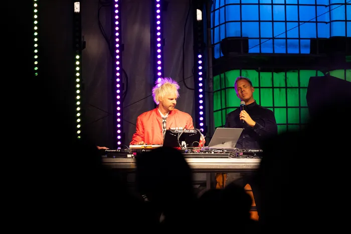Ein DJ mit blonden, stacheligen Haaren in einer roten Jacke bedient Mischpulte neben einem Mann mit Mikrofon vor bunten LED-Lichtstreifen.