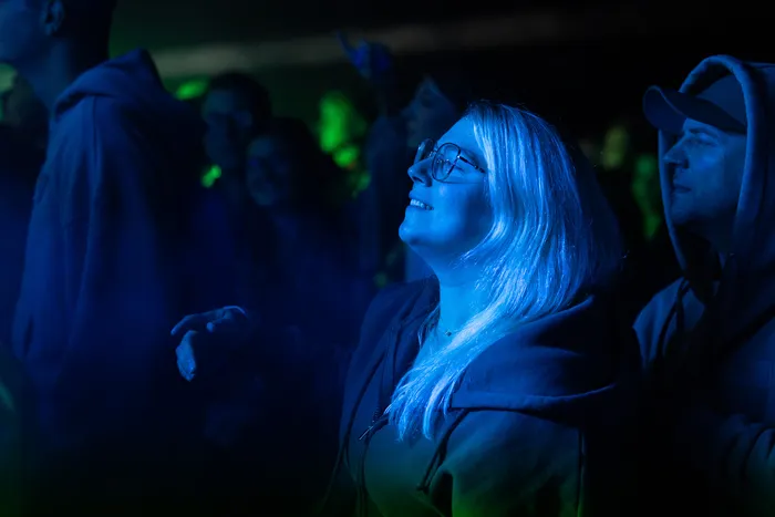 Eine blondhaarige Frau mit Brille blickt unter blauem Licht nach oben, umgeben von einer Menschenmenge.