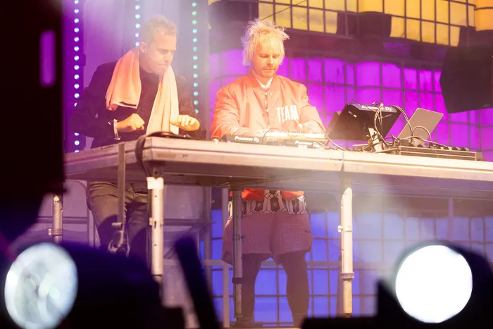 Zwei DJs bedienen DJ-Equipment an einem Pult vor einer lila und gelb beleuchteten Bühnenkulisse.