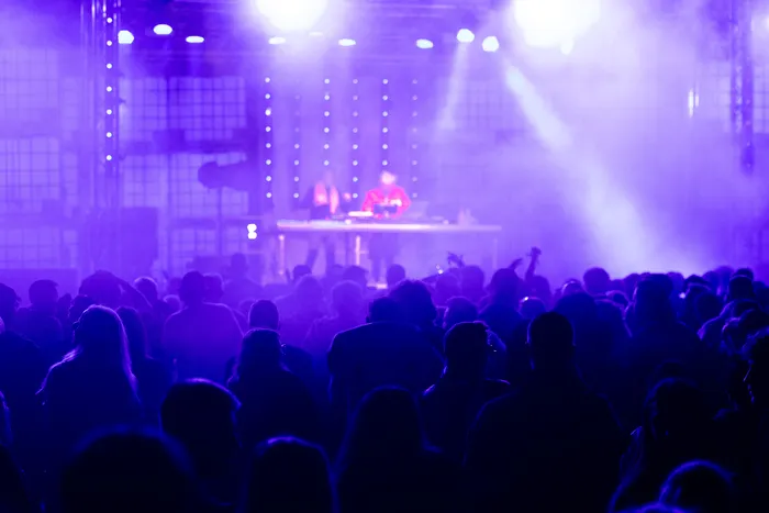 Silhouetten einer Menschenmenge vor einer lila beleuchteten Bühne mit zwei Performern und Bühnennebel.