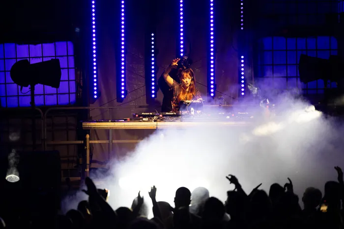 DJ Lari Luke performt an einem Turntable-Pult vor blauen Lichtstreifen und dichtem Bühnennebel vor einem silhouettierten Publikum.