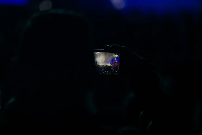 Silhouette einer Person, die mit einem Smartphone eine beleuchtete Bühne und eine Menschenmenge bei einem Event fotografiert.