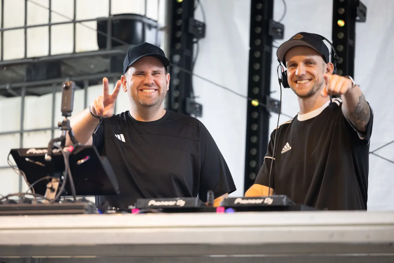 Die DJs Mush-T lächeln hinter ihrem Equipment; einer macht ein Peace-Zeichen, der andere zeigt auf die Kamera.