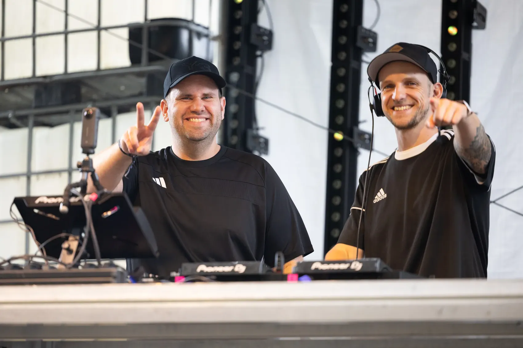 Die DJs Mush-T lächeln hinter ihrem Equipment; einer macht ein Peace-Zeichen, der andere zeigt auf die Kamera.