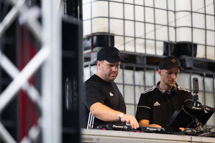 Zwei Männer in schwarzen T-Shirts und Kappen bedienen DJ-Equipment auf einer Bühne vor großen weißen Containern.