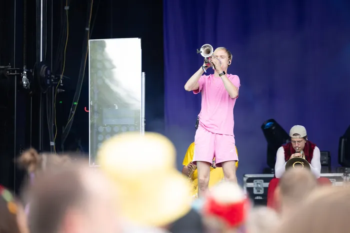Ein Mann in rosa Kleidung spricht in ein Megafon auf einer Bühne, im Hintergrund ist ein Musiker mit einem Blechblasinstrument zu sehen.