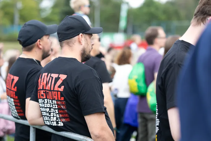 Männer in schwarzen T-Shirts mit dem roten „BAUTZ FESTIVAL“-Schriftzug stehen an einer Metallabsperrung vor einer Menschenmenge.