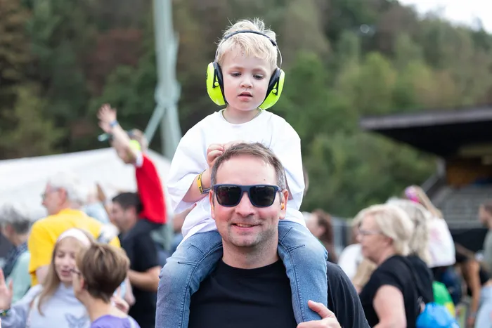 Ein kleiner Junge mit neongrünen Kopfhörern sitzt auf den Schultern eines Mannes mit Sonnenbrille bei einem Open-Air-Event.