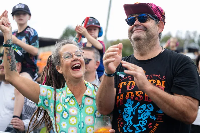 Eine lachende Frau mit Dreadlocks und Brille hebt den Arm, während ein Mann in einem schwarzen T-Shirt daneben klatscht.