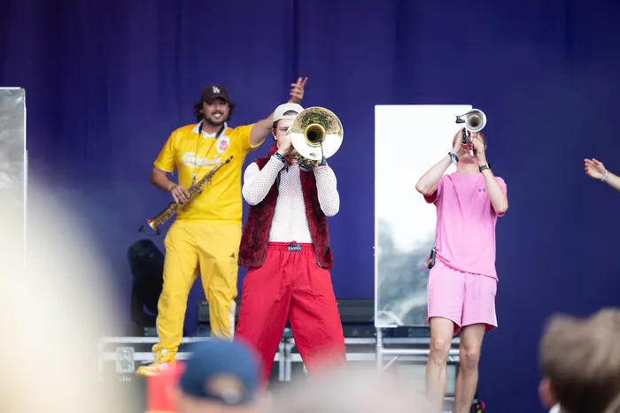 Musiker in gelber, roter und rosa Kleidung spielen Saxophon, Tuba und Trompete auf einer Bühne vor blauem Hintergrund.